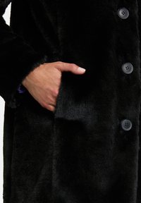 Black faux fur coat with a smooth texture, features a side pocket, and four buttons in a matte finish. Close-up of hand in pocket.