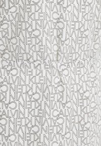 Tissu blanc avec un motif récurrent en texte noir "CALVIN KLEIN", comportant une poche avant et des coutures visibles.