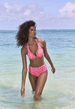 Frau mit lockigem Haar, die einen pink gemusterten Bikini trägt und bis zur Taille in klarem Ozeanwasser watet, unter einem blauen Himmel mit verstreuten Wolken.