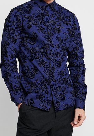 Mann trägt ein tailliertes blaues Hemd mit schwarzen Blumenmustern und schwarze Hose, Hände ruhen nahe den Taschen.