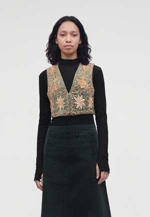 Une femme porte un top noir et une longue jupe foncée avec un gilet orné de broderies verdes et dorées sur un fond uni.