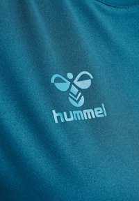 Teal idrottströja med en mjuk, slät textur, med en ljusblå "hummel"-logotyp och ett abstrakt mönster tryckt på framsidan.
