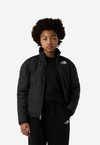 Μαύρο μπουφάν puffer με ψηλό γιακά και κλείσιμο με φερμουάρ, με λογότυπο στο αριστερό στήθος. Φορεμένο πάνω από μαύρο πουλόβερ και παντελόνι φόρμας.