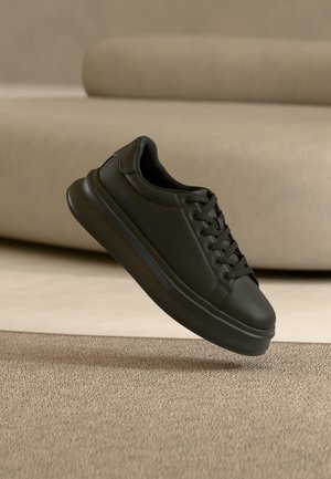 Pantof sport din piele neagră, cu talpă groasă și șireturi, plutind deasupra unui covor bej într-un living cu tonuri neutre.