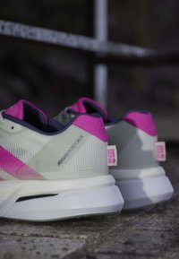 Par lysegrå og pink løbesko med hvide såler placeret på en stengulv, der viser hæl- og sidedetaljer.