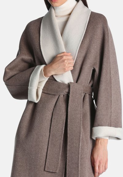 Femme portant un manteau en laine marron ceinturé avec un large col châle blanc et des détails aux poignets, par-dessus un pull à col roulé blanc.