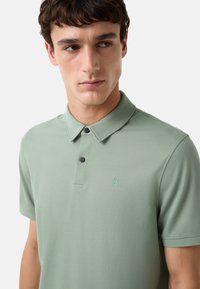 Polo T-shirt i lysegrønt stof, har korte ærmer, en skjortekrave med to sorte knapper og et lille logo i matchende farve på brystet.
