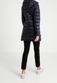 Navy quilted puffer coat with a hood, featuring a fitted silhouette and side pockets, paired with black pants and white sneakers.