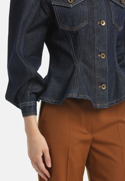 Giacca di jeans scura con chiusura a bottoni, tasche sul petto e maniche a palloncino; abbinata a pantaloni marroni su misura. Accenti in metallo dorato evidenti.