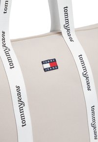 Μπεζ τσάντα tote με λευκές λουρίδες, με το λογότυπο "TOMMY JEANS" σε ναυτικό μπλε και κόκκινο στο μπροστινό μέρος. Λεία υφή, κλείσιμο με φερμουάρ.