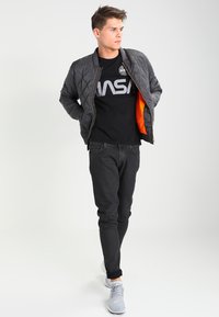 Chaqueta bomber acolchada gris con forro naranja, usada sobre una camiseta negra de la NASA, combinada con jeans negros y zapatillas gris.