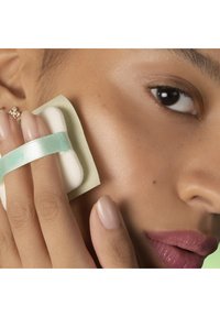 Éponge cosmétique avec une sangle vert menthe, reposant sur la peau. Forme douce et ronde, texture blanche, tenue entre les doigts près des lèvres et de la joue.