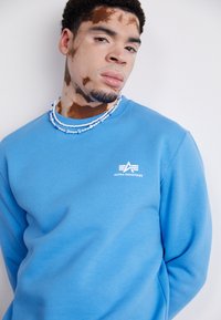 Felpa blu con collo a giro e un piccolo logo bianco sul petto. Indossata con una collana di perline multicolore. Texture del tessuto liscia.
