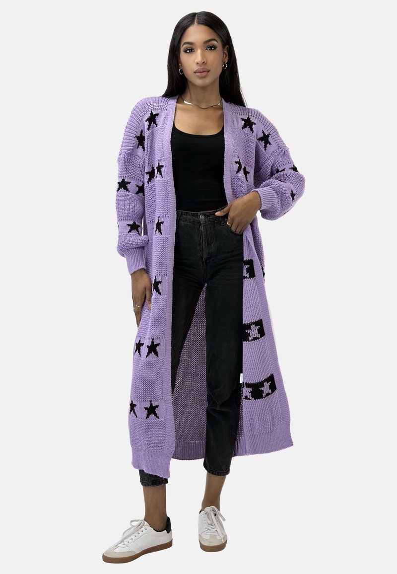Fioletowy dziergany kardigan z czarnymi wzorami gwiazd, długimi rękawami, otwartym przodem i swobodnym krojem, zestawiony z ciemnymi dżinsami i białymi trampkami.