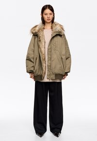 Parka acolchada en verde oliva con collar de piel sintética color beige desmontable, cierre con cremallera y dos bolsillos laterales, combinado con pantalones de pierna ancha negros.