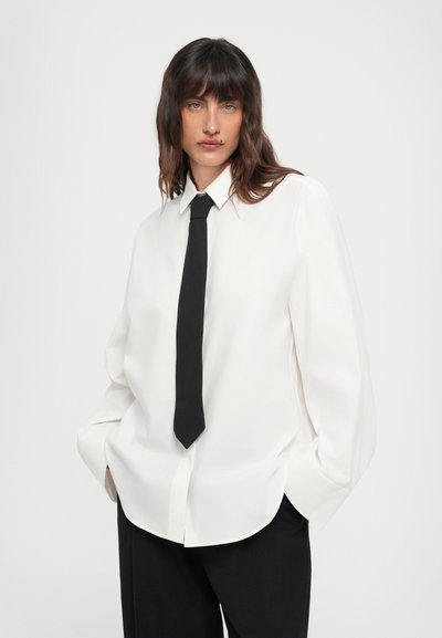 Camicia bianca a maniche lunghe con colletto a punta, abbinata a una cravatta nera slim. La camicia ha una vestibilità rilassata con orlo arrotondato. Tessuto testurizzato.