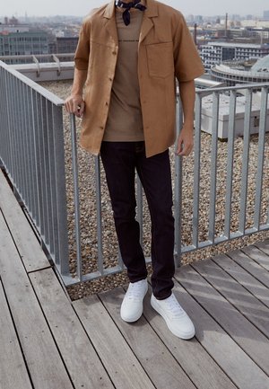 Osoba stojąca na tarasie na dachu, ubrana w brązową kurtkę z krótkim rękawem, beżową koszulę, ciemne dżinsy, białe trampki i czarną apaszkę na szyi.