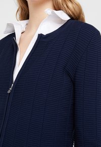 Cazadora navy de punto con cremallera, con textura de canalé y una camiseta blanca con cuello debajo. Detalle de cremallera plateada visible en el cuello.