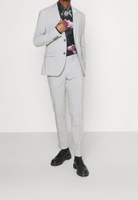 Traje gris claro de corte ajustado, con una camisa floral negra debajo. Zapatos negros con costuras decorativas completan el look.