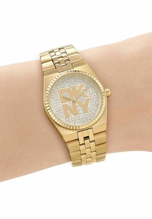 Montre-bracelet DKNY ton or avec un cadran incrusté de cristaux et un logo DKNY audacieux, portée sur un poignet à la peau claire.