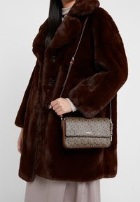 Žena v tmavě hnědém kabátu z umělé kožešiny drží hnědou crossbody tašku s monogramem DKNY, která má kožený popruh a zlatý řetízek.