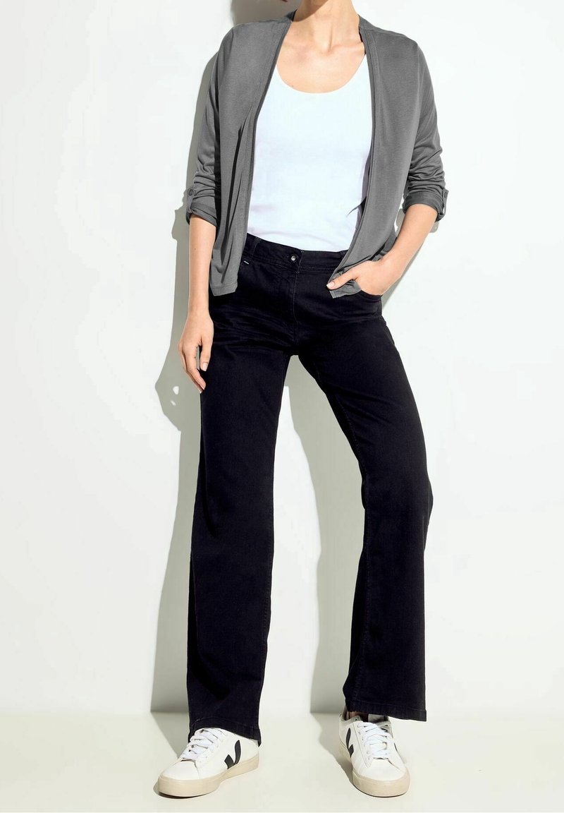 Femme portant un cardigan gris ouvert, un débardeur blanc, un pantalon large noir et des baskets blanches, debout contre un mur blanc.