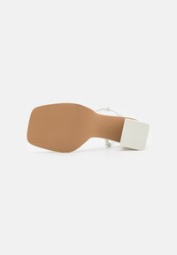 Sandale blanche à talon haut avec un talon carré et une semelle lisse, couleur marron clair. Dispose de fines lanières pour un design délicat.