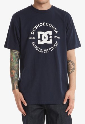 Námořnická modrá bavlněná tričko s bílou grafikou a textem "DCSHOECOUSA" a "ATLETICKÁ SKATEBOARDOVÁ OBUV A OBLEČENÍ". Krátké rukávy, kulatý výstřih.