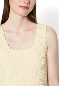 Top de alças de cor creme claro com um padrão de crochê texturizado, apresentando uma gola em forma de concha e design sem mangas.