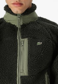 Ciemnozielona polarowa kurtka z teksturowanym, puszystym wykończeniem, zapinana na zamek z przodu i kontrastowymi oliwkowymi detalami. Logo Lacoste na klatce piersiowej.