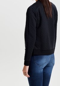 Veste bomber matelassée noire avec une taille et des poignets ajustés, présentée avec un jean bleu. Elle possède un motif texturé et un col rond.