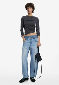 Graues, kurz geschnittenes Langarmshirt mit "Desigual"-Text, hellblaue weite Jeans, schwarze Handtasche und schwarze Schuhe mit grünen Schnürsenkeln.