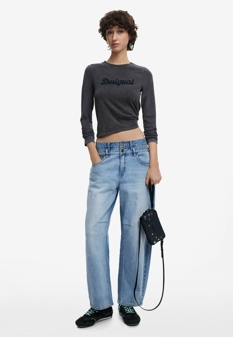 Graues, kurz geschnittenes Langarmshirt mit "Desigual"-Text, hellblaue weite Jeans, schwarze Handtasche und schwarze Schuhe mit grünen Schnürsenkeln.