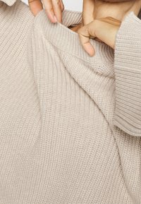 Maglione beige chiaro lavorato a maglia con colletto pieghevole a coste. La texture appare morbida e spessa, evidenziando il suo design accogliente e i dettagli del tessuto.