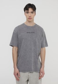 Maglietta grigia oversize con aspetto scolorito, con la scritta "WEST COAST" ricamata con filo scuro sul petto. Materiale in cotone morbido.