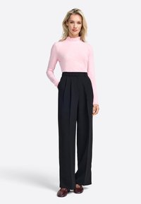 Roze langetees met gerimpelde kraag, gecombineerd met hoge zwarte geplooide broek en bordeauxrode schoenen, tegen een neutrale achtergrond.