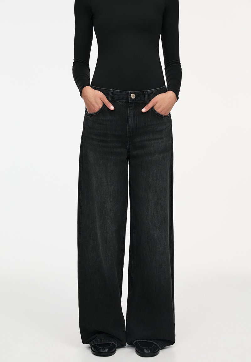 Femme portant un jean noir à jambes larges et un haut noir ajusté à manches longues, les mains dans les poches, debout sur un fond blanc.