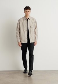 Light beige button-up shirt with a chest pocket, paired with black trousers and black shoes; smooth fabric, straight-cut design.
