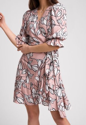 Donna che indossa un abito rosa a portafoglio con grande stampa floreale bianca e orlo arricciato, in piedi davanti a uno sfondo semplice.