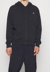 Man i svart zip-up hoodie med en liten vit Jordan-logga på bröstet och svarta byxor, stående mot en neutral bakgrund.