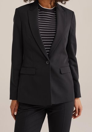 Vrouw draagt een aansluitende zwarte blazer met één knoop, klepzakken en een zwart-wit gestreept shirt eronder.