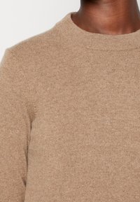 Pull tricoté couleur sable avec un col rond et une texture côtelée, possédant des manches longues et une finition douce et lisse.