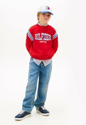 Chłopiec w czerwonej bluzie Hilfiger, niebieskiej koszuli w paski, jasnoniebieskich dżinsach, granatowych trampkach i białej czapce na białym tle.