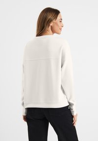 Jasnobeżowy sweatshirt o luźnym kroju, z długimi rękawami, ściągaczami na mankietach oraz poziomym szwem na plecach. Nosi się go z czarnymi spodniami.
