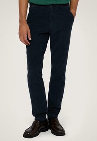 Námořnické bavlněné kalhoty s úzkým střihem, obsahující přední kapsy a hladkou texturu, spárované s tmavě hnědými mokasíny.