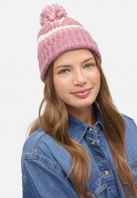Gorro de punto rosa con una franja blanca, que presenta un pompón en la parte superior y una textura acanalada. Combinado con una camisa vaquera.