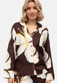 Conjunto de pijama marrón con estampado floral que presenta grandes flores blancas y amarillas, con botones en el frente y mangas largas con ribete amarillo en contraste.