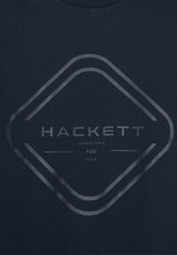 Námornícky modré tričko s veľkým kruhovým logom, na ktorom sú v tučných písmenách napísané slová "HACKETT" a šikmé akcenty. Mäkký bavlnený materiál, krátke rukávy.