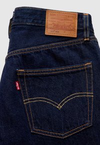 Τσέπη στο πίσω μέρος σκούρου μπλε τζιν με κίτρινη ραφή, κόκκινη υφασμάτινη ετικέτα Levi's και δερμάτινο ταμπελάκι μάρκας tan Levi Strauss & Co.