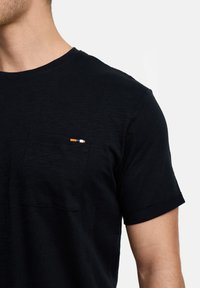 Sort t-shirt med korte ærmer, rund halsudskæring og en plysdolk lomme med en lille stribet detalje i orange, hvid og blå.